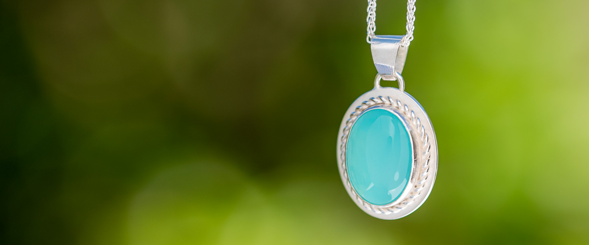 A photo of a silver pendant with large aqua coloured cabochon gemstone hanging on a silver chain in front of out of focus green foliage in the background