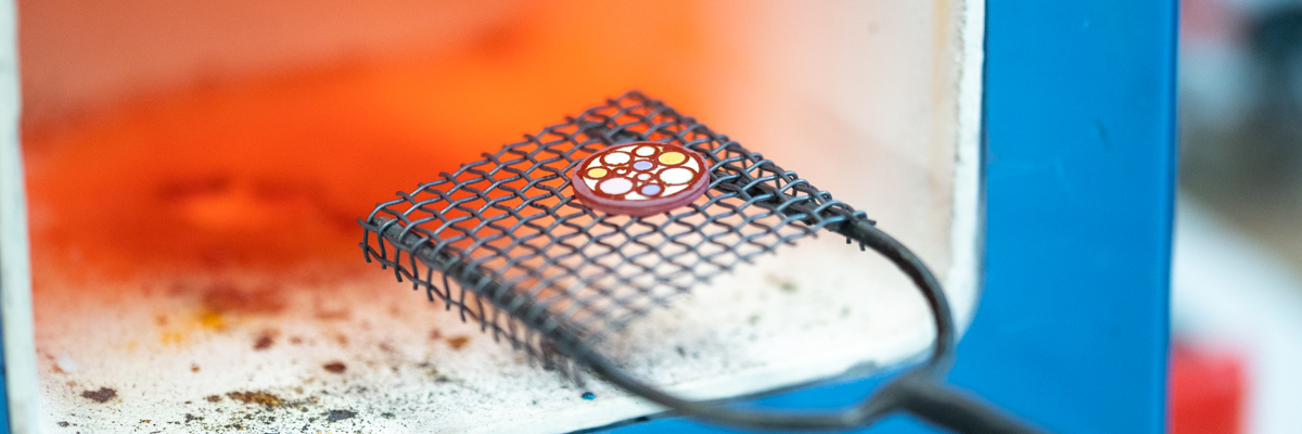 Enamelled jewellery piece being taken out of a hot glowing jewellery kiln