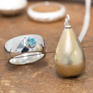 Hollow form silver jewellery on a jewellery bench including rings and a teardrop shaped textured pendant with gold embellishment.