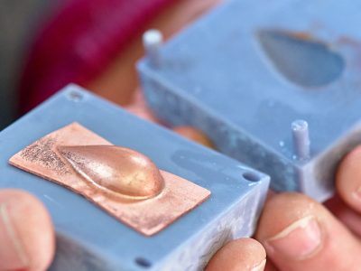 Close up photo of a small copper sheet domed into a teardrop shape next to a teardrop shaped die.